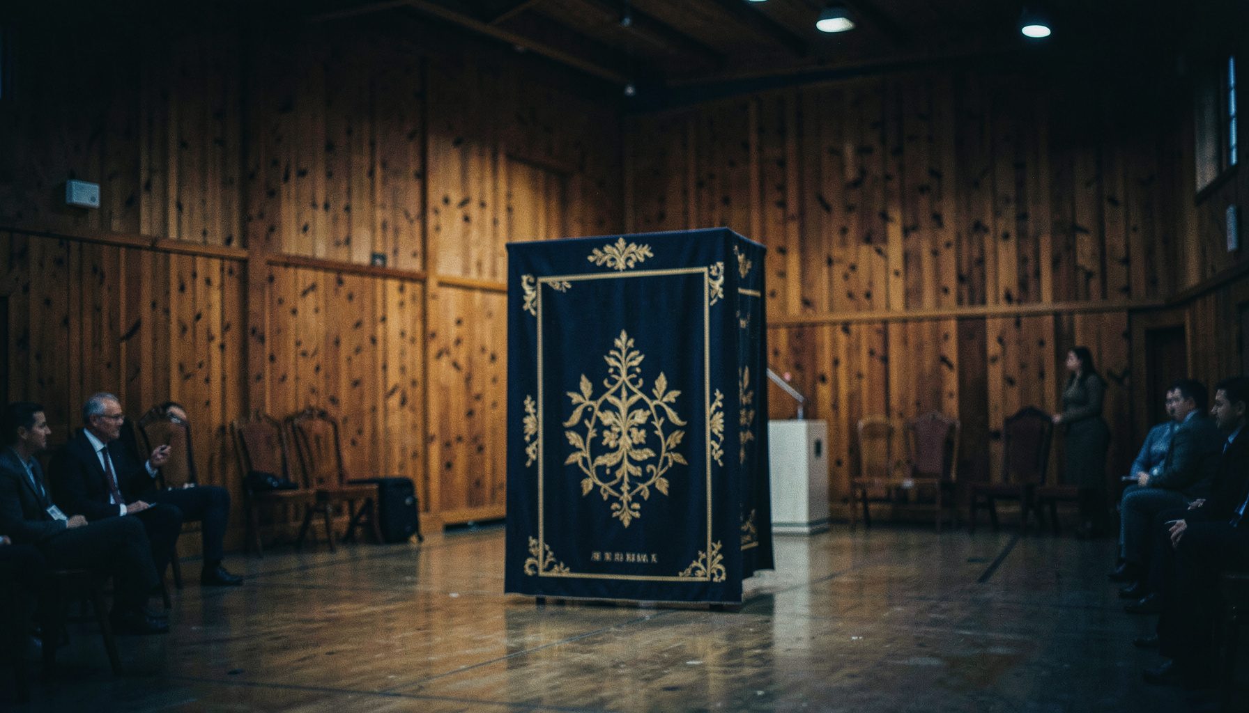 En högtidlig ceremoni inomhus i en rustik mässlokal med varma träpaneler och dämpad belysning. Ett standar med mörkblå och guldfärgade detaljer står i centrum.